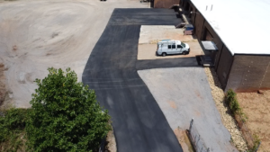 A newly paved asphalt driveway leads to a commercial building with a white van parked in a designated space, surrounded by gravel and some vegetation.