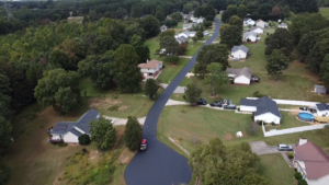 A newly paved road runs through a suburban neighborhood with houses, trees, and lawns visible on both sides.