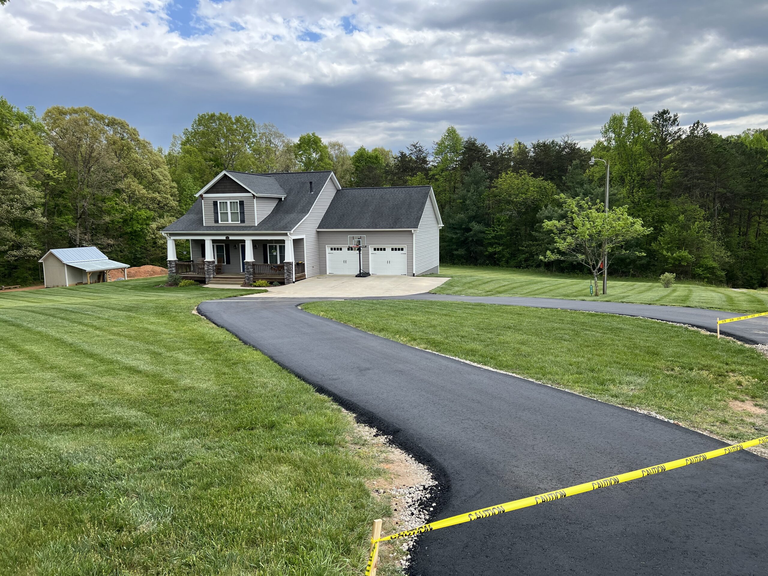 A newly paved asphalt driveway leads to a gray two-story house with a porch and double garage, surrounded by green grass and trees; caution tape blocks off the driveway's entrance.