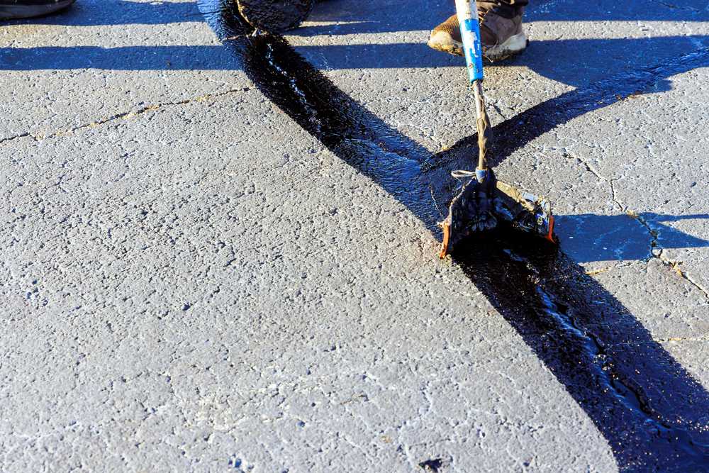 A person seals a crack in asphalt pavement with black tar using a long-handled applicator tool.