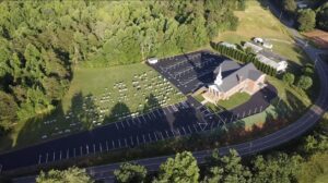 Aerial view of a church with adjacent cemetery, parking lot, and surrounding trees, bordered by a curved road.