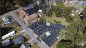 Aerial view of a parking lot next to a brick building, surrounded by trees, houses, and roads in a residential neighborhood.