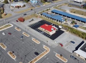 Aerial view of a small fast-food restaurant with a red roof in the center, surrounded by large mostly empty parking lots and nearby roads.