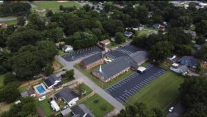 Aerial view of a large building with parking lots, surrounded by trees, houses, and green spaces in a suburban neighborhood.