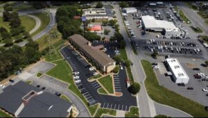 Aerial view of a commercial area with multiple buildings, parking lots, and cars, bordered by roads and greenery.