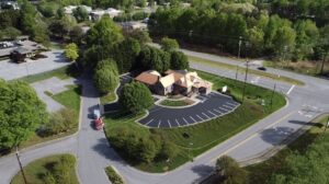 Aerial view of a restaurant with a red roof, surrounded by trees and an empty parking lot, located at the intersection of two roads.