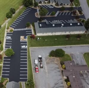 Aerial view of a newly paved parking lot with white painted lines, adjacent to a building and an older, faded parking area with maintenance vehicles present.