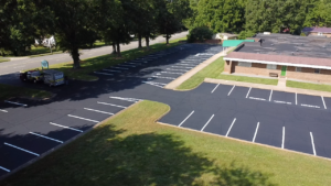 A freshly paved and striped parking lot with marked spaces beside a brick building, bordered by grass and trees.