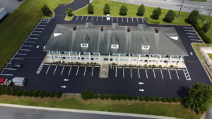 Aerial view of a two-story motel with a gray roof, surrounded by a freshly paved, mostly empty parking lot with marked spaces and landscaped greenery.