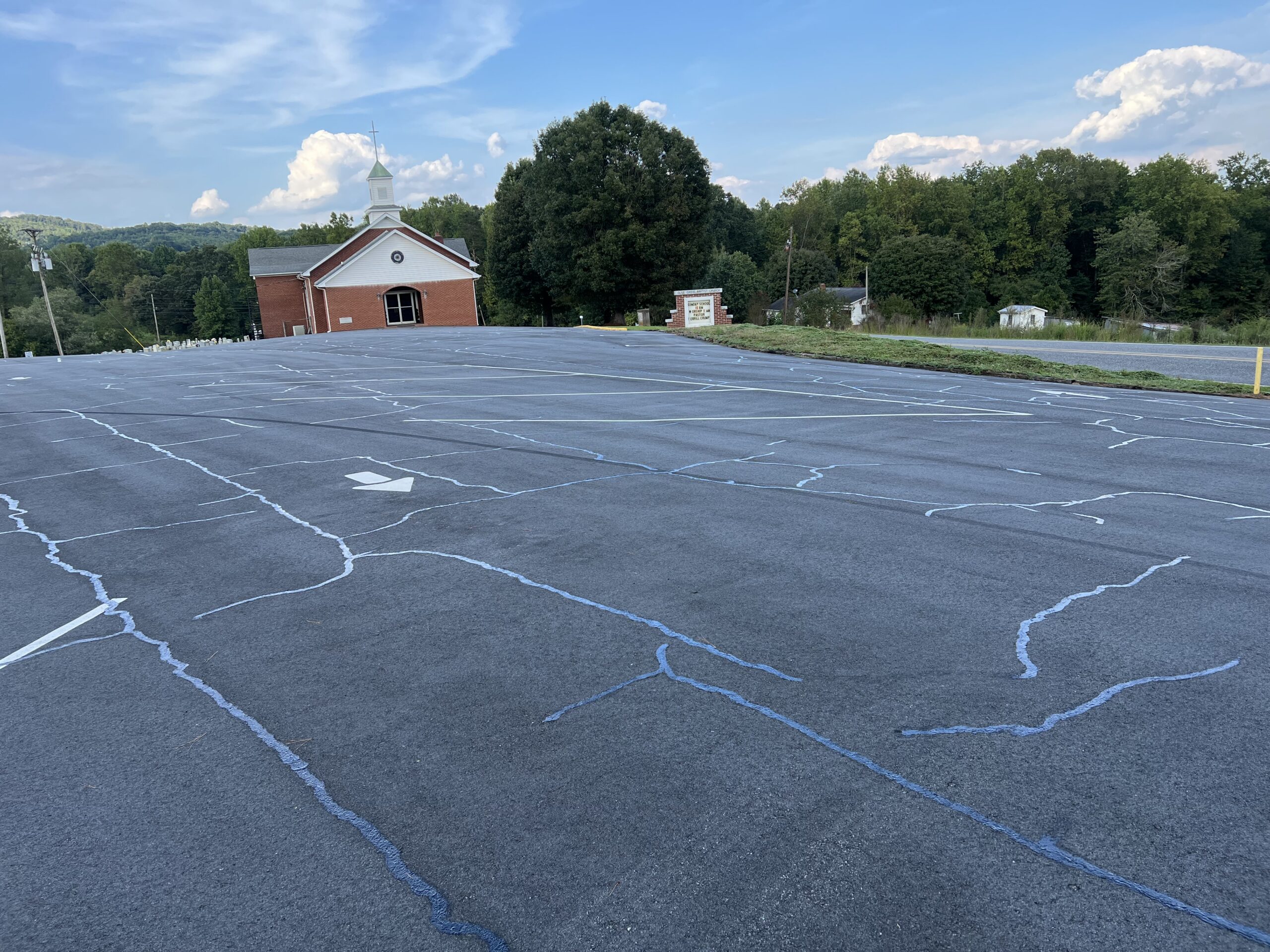 Large empty parking lot with visible crack repairs, white directional arrow, and a small red brick church building in the background surrounded by trees under a partly cloudy sky.