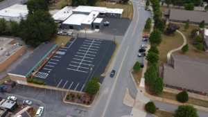 Aerial view of a freshly paved parking lot with white lines, adjacent buildings, trees, and several cars parked along nearby roads.