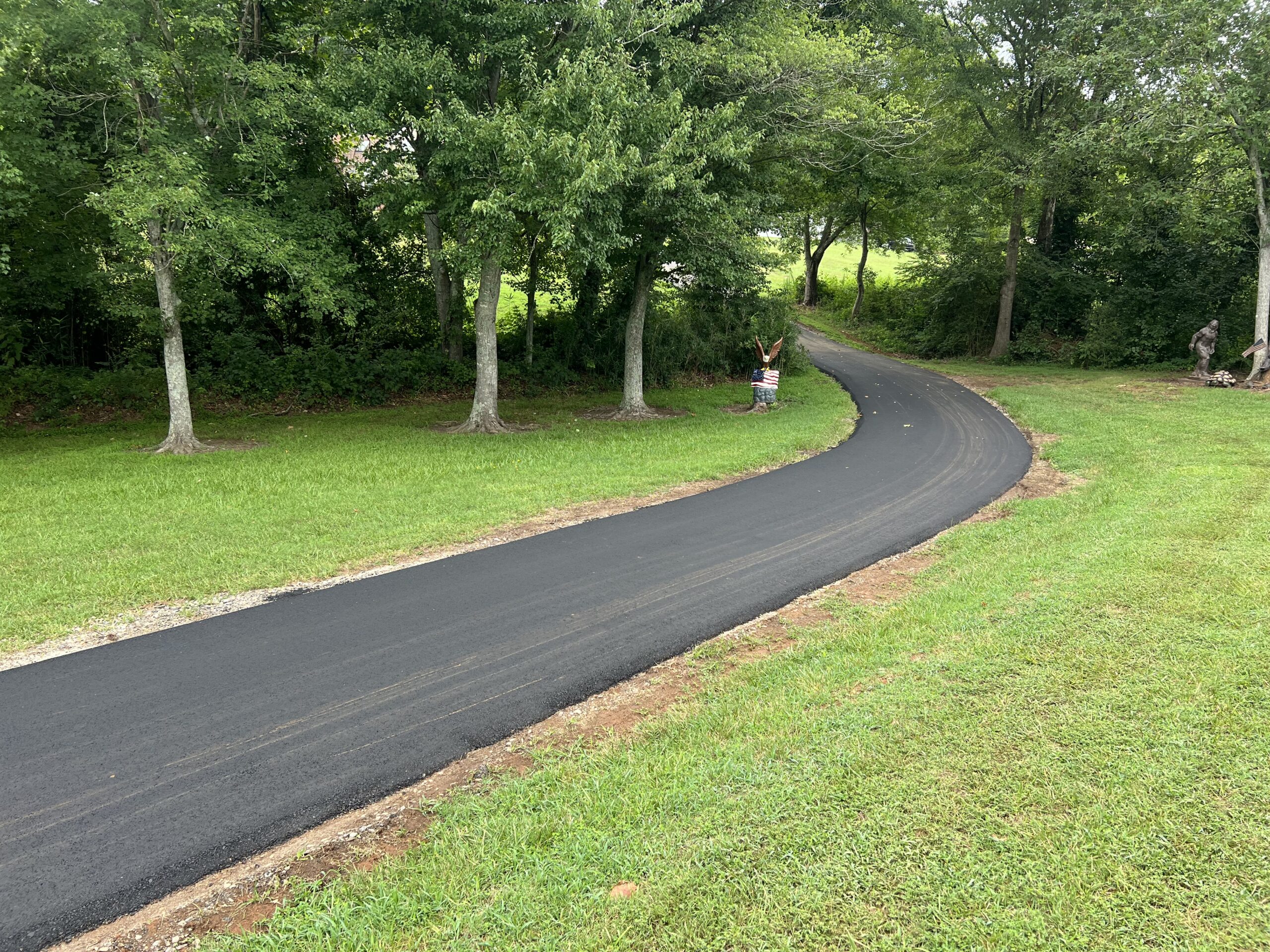 A newly paved asphalt path curves through a grassy area with trees, while a person stands near the side of the path with arms raised.