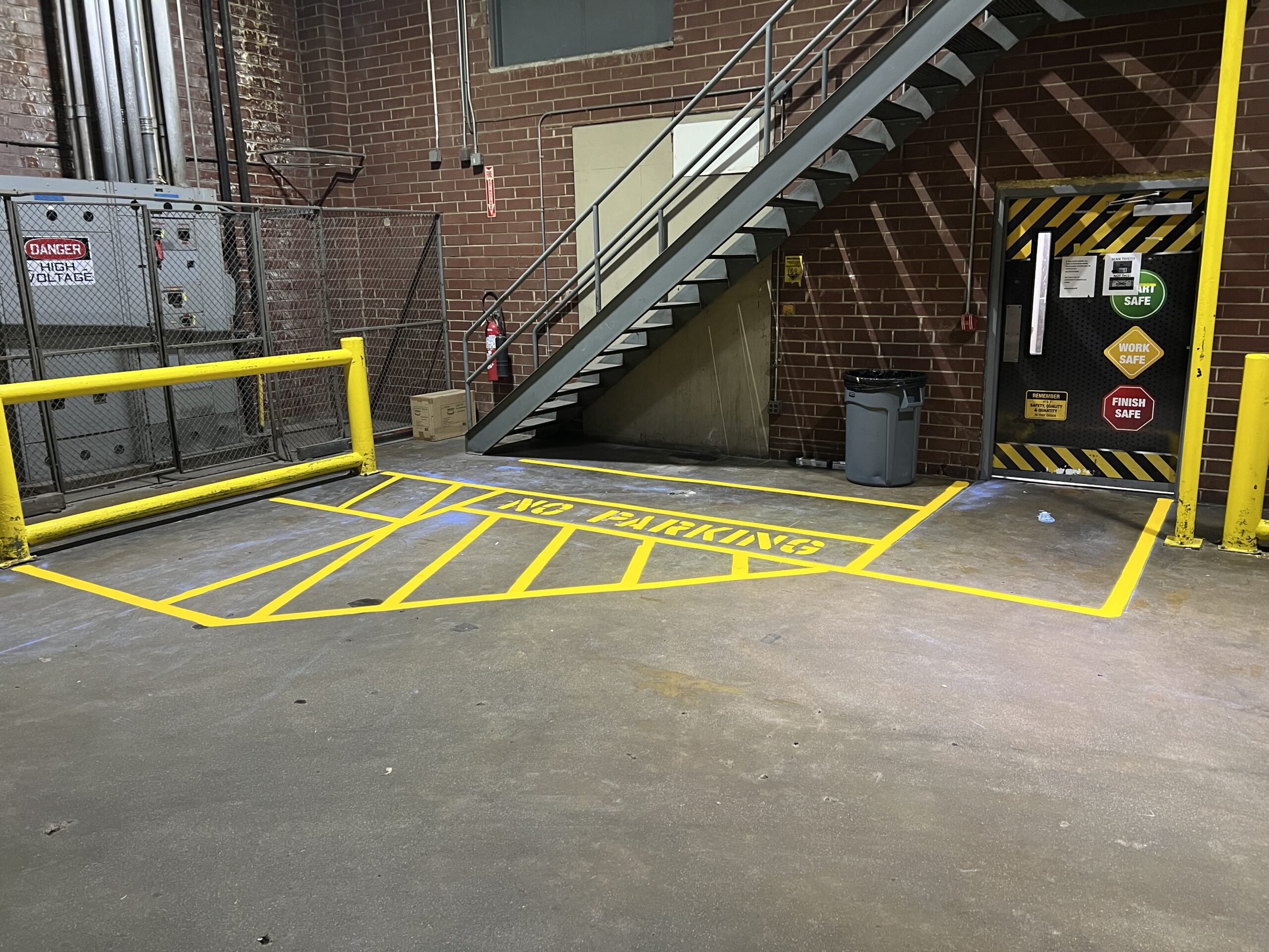 Yellow lines on a concrete floor mark a NO PARKING zone near a metal staircase and a door in an industrial area with brick walls and safety signs.